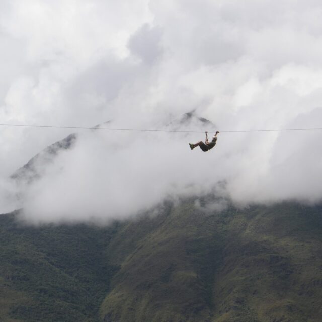 Someone high in the sky zip lining across a valley before the mountains and clouds.